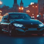 A sleek BMW M3 parked at night on a city street with bright streetlights casting reflections off its glossy black paint.