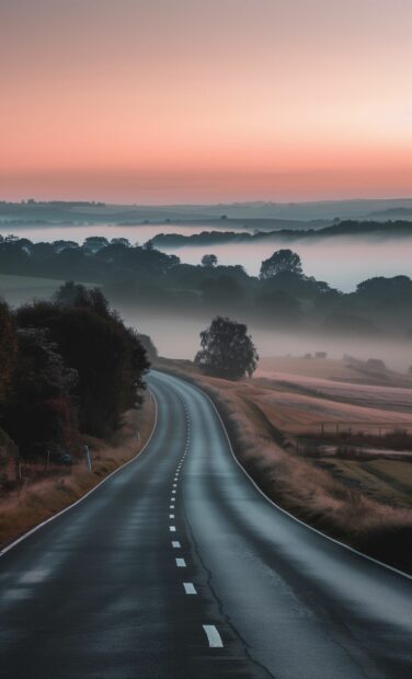 A country road at dawn with soft pastel colors in the sky and morning mist hovering over the fields.