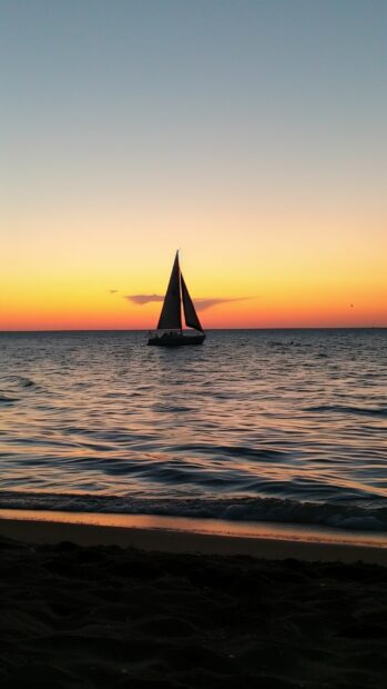A serene beach at sunset with a sailboat on the horizon, Ocean Aesthetic wallpaper.