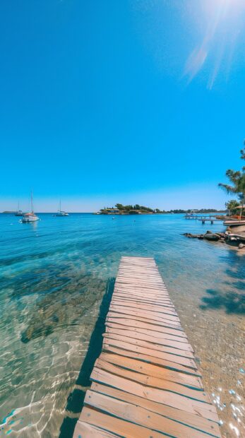 A preppy beach iPhone wallpaper with a picturesque boardwalk, sailboats in the distance, and clear blue skies.