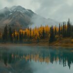 A serene mountain background HD with a crystal clear lake in the foreground and autumn colored trees.