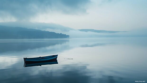 A tranquil morning mist over a calm lake with a lone fishing boat and distant mountains.