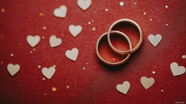 Happy Valentine Day Desktop Background with two gold wedding rings intertwined on a velvet red surface, surrounded by tiny heart confetti, elegant and romantic.