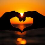 Love Wallpaper with a close up of a couple's hands forming a heart shape against the backdrop of a sunset, creating a warm and loving mood.