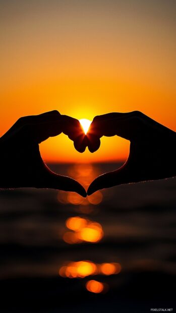 Love Wallpaper with a close up of a couple hands forming a heart shape against the backdrop of a sunset, creating a warm and loving mood.